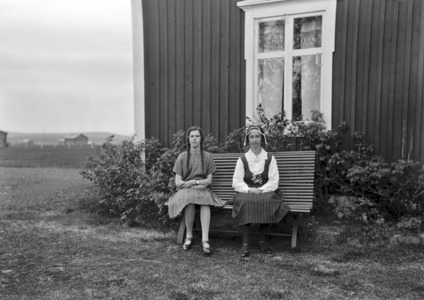 Svartvit bild från 1920-talet, av två yngre kvinnor på en bänk på gräset utanför huset.