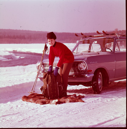 Äldre bild på leende kvinna som packar en ryggsäck utanför bilen på vintern. På takräcket två par skidor.