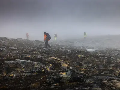 'Fyra arkeologer med ryggsäckar går i dimman i ett platt och klippigt bergslandskap i Norrbottensfjällen.'