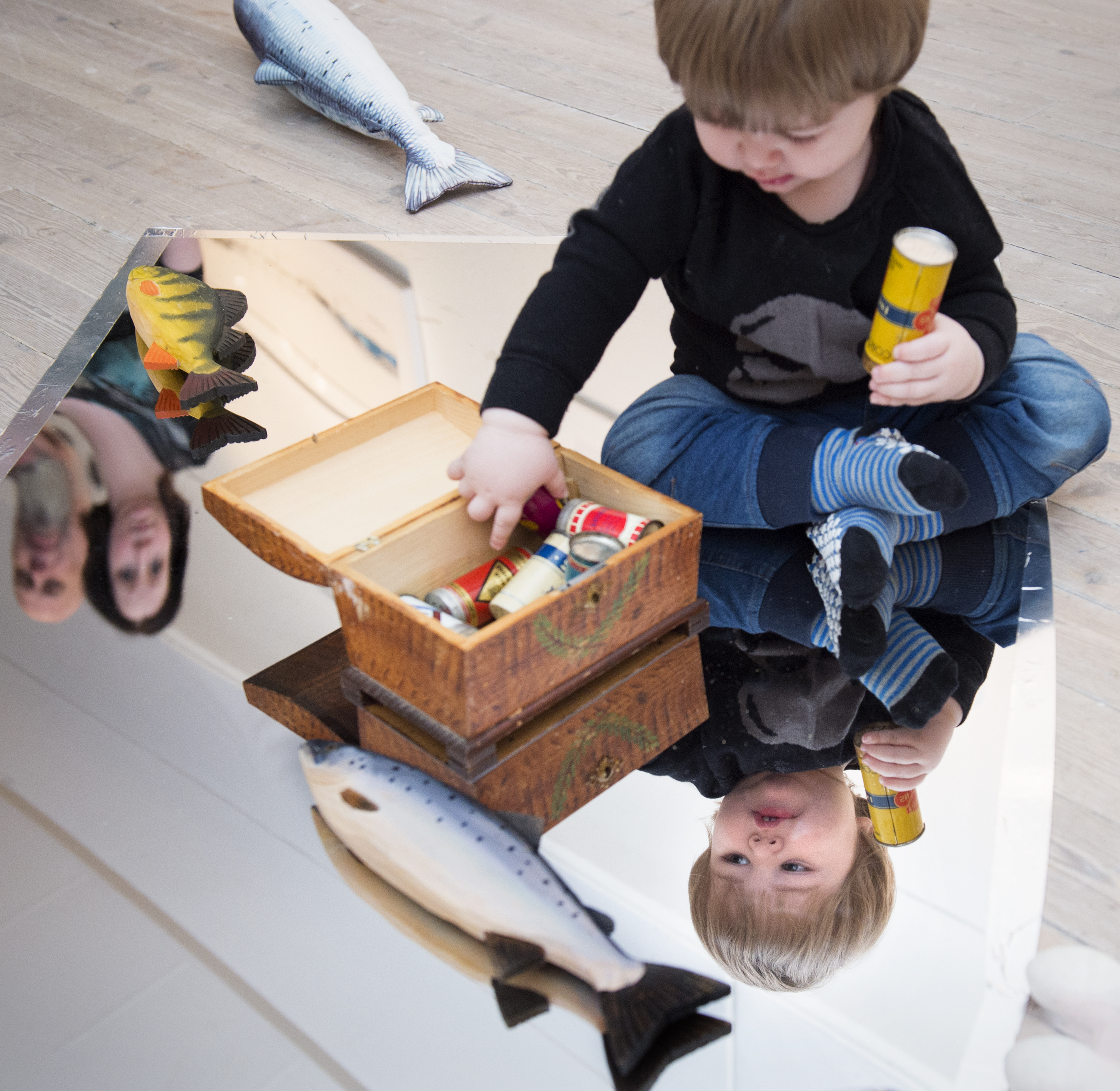 'Liten pojke som leker med en ask. Han sitter på en spegel föreställande en älv. Två fiskar av trä ligger på sidan om honom.'