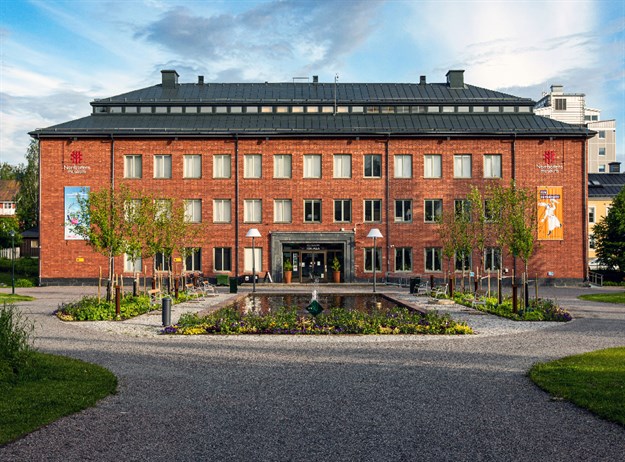 'Norrbottens museum i Hermelinsparken i Luleå. Framför byggnaden finns en damm, träd och blommor. Det är sommar.'