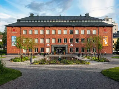 'Norrbottens museum i Hermelinsparken i Luleå. Framför byggnaden finns en damm, träd och blommor. Det är sommar.'