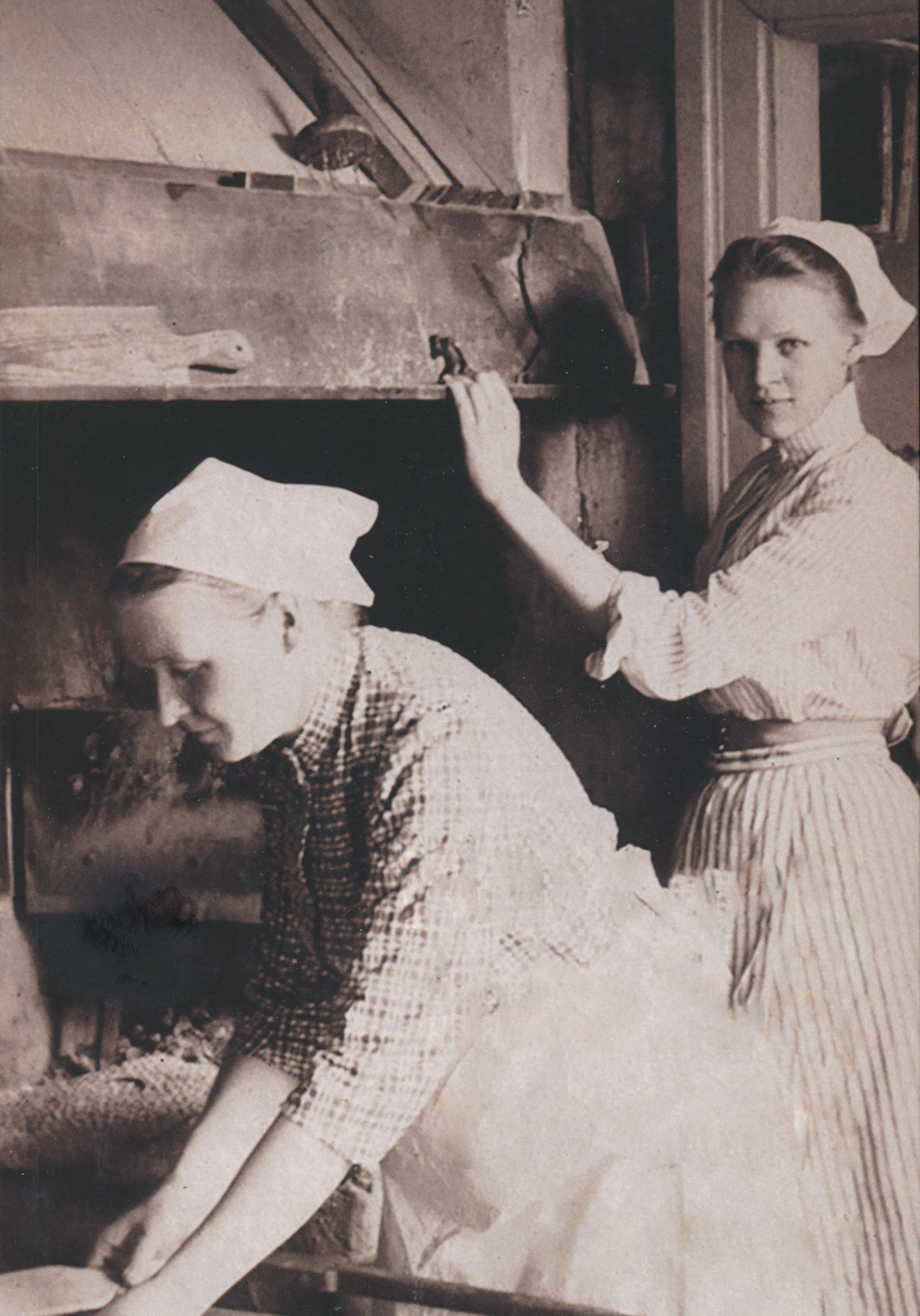 Äldre bild av två kvinnor som står vid en spis och lagar mat.