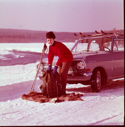 Kvinna med skidutrustning som står på isväg framför en bil. 1960-tal.
