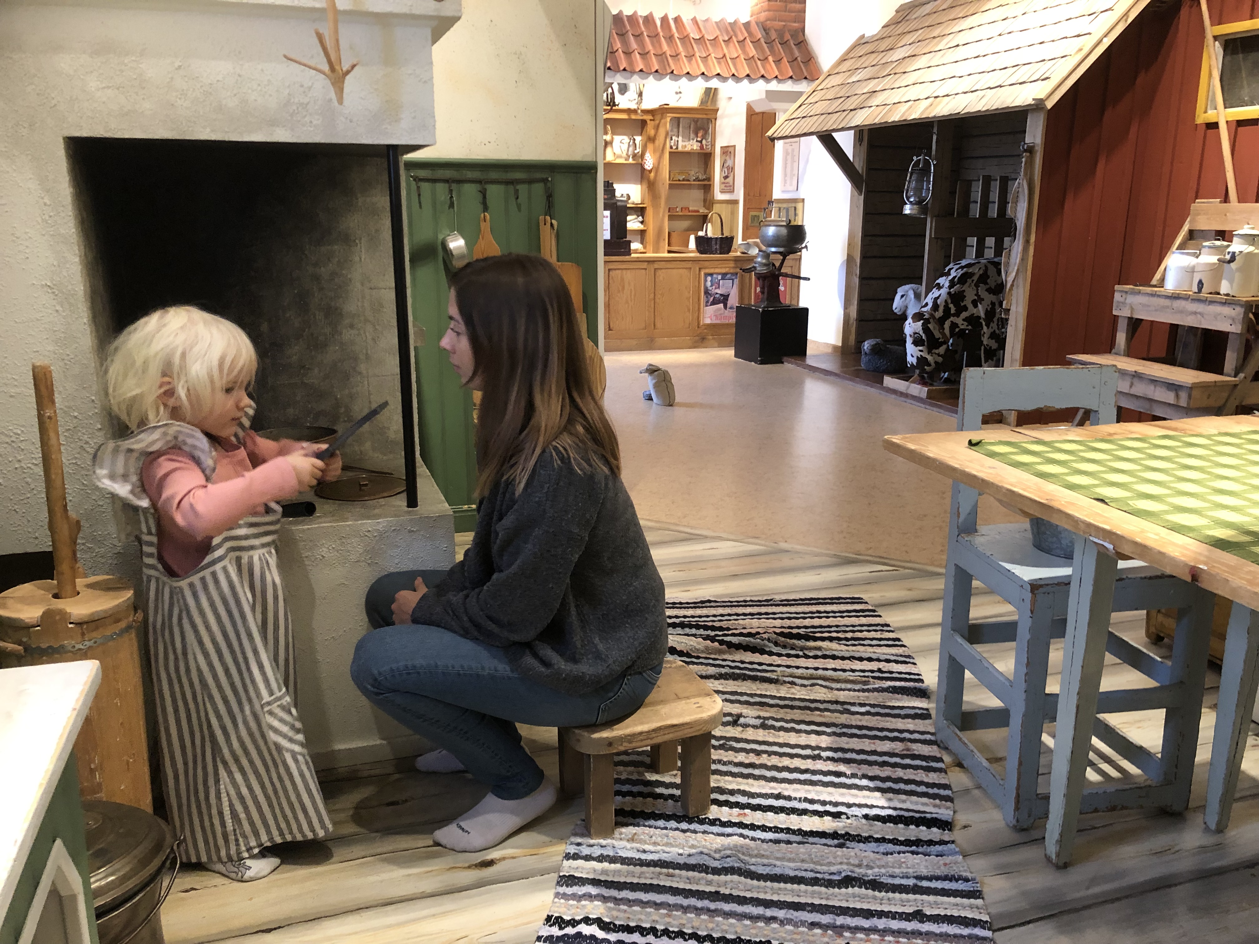 'Barn och förälder som leker vid den öppna spisen i köket i lekmiljön Förr i tiden. I bakgrunden skymtar handelsboden och ladugården.'