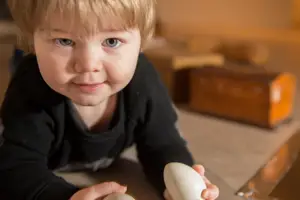 En liten pojke som ligger på mage och tittar in i kameran. I händerna håller han två ägg av trä.