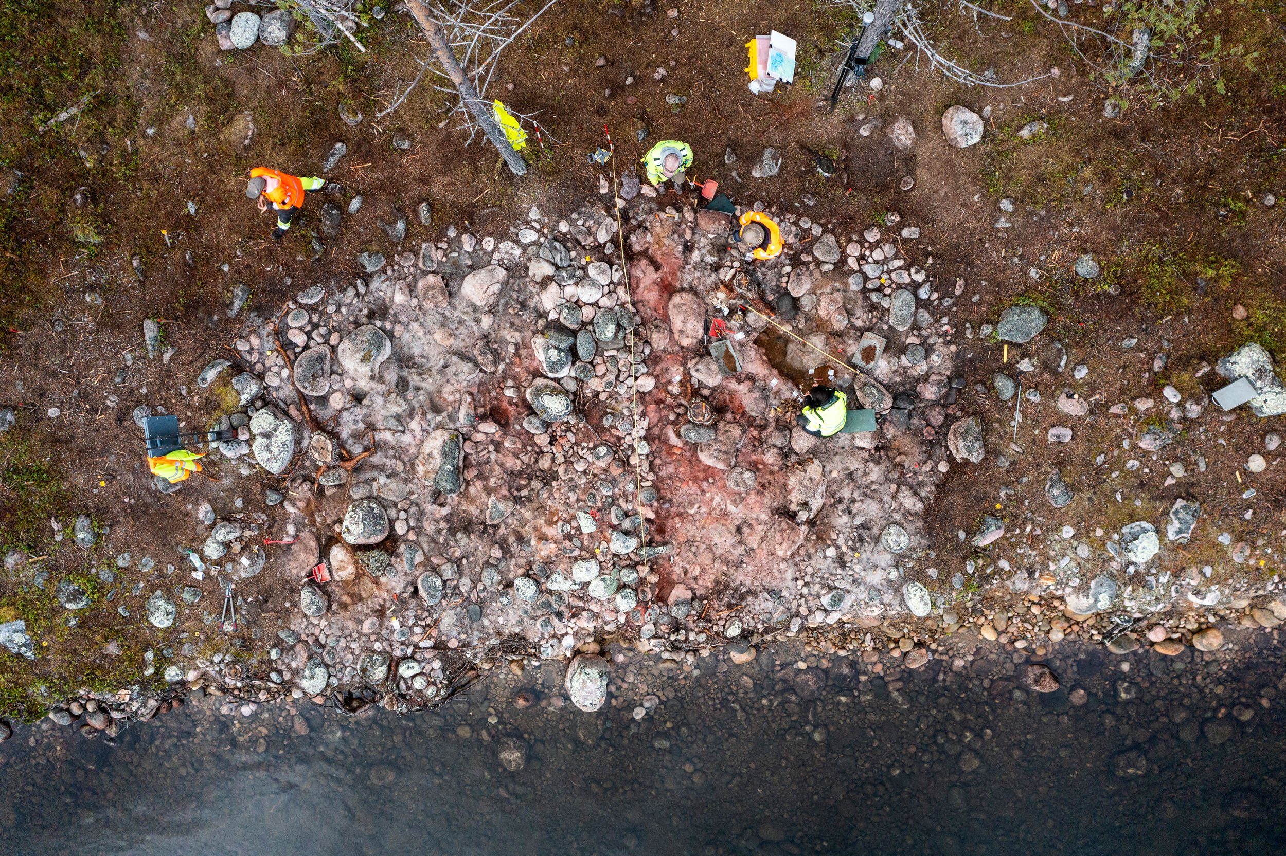 Vy över stenåldersgrav i Liggá. Fem arkeologer gör en arkeologisk utgrävning vid graven. På platsen finns det stenar.