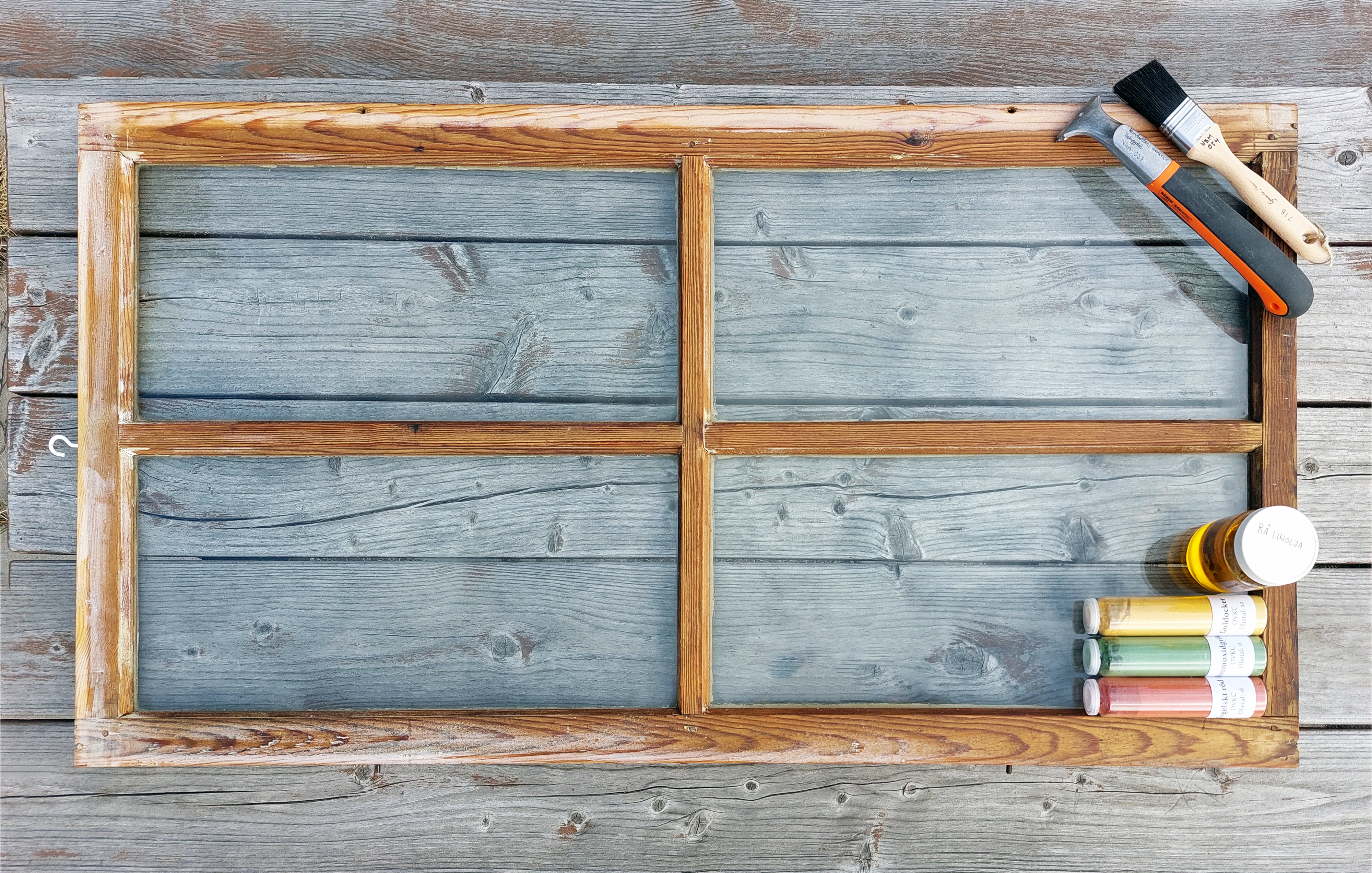 'Gammalt fönster som ligger på ett bord. På sidan ligger färger, en pensel och en skrapa att ta bort färg med.'