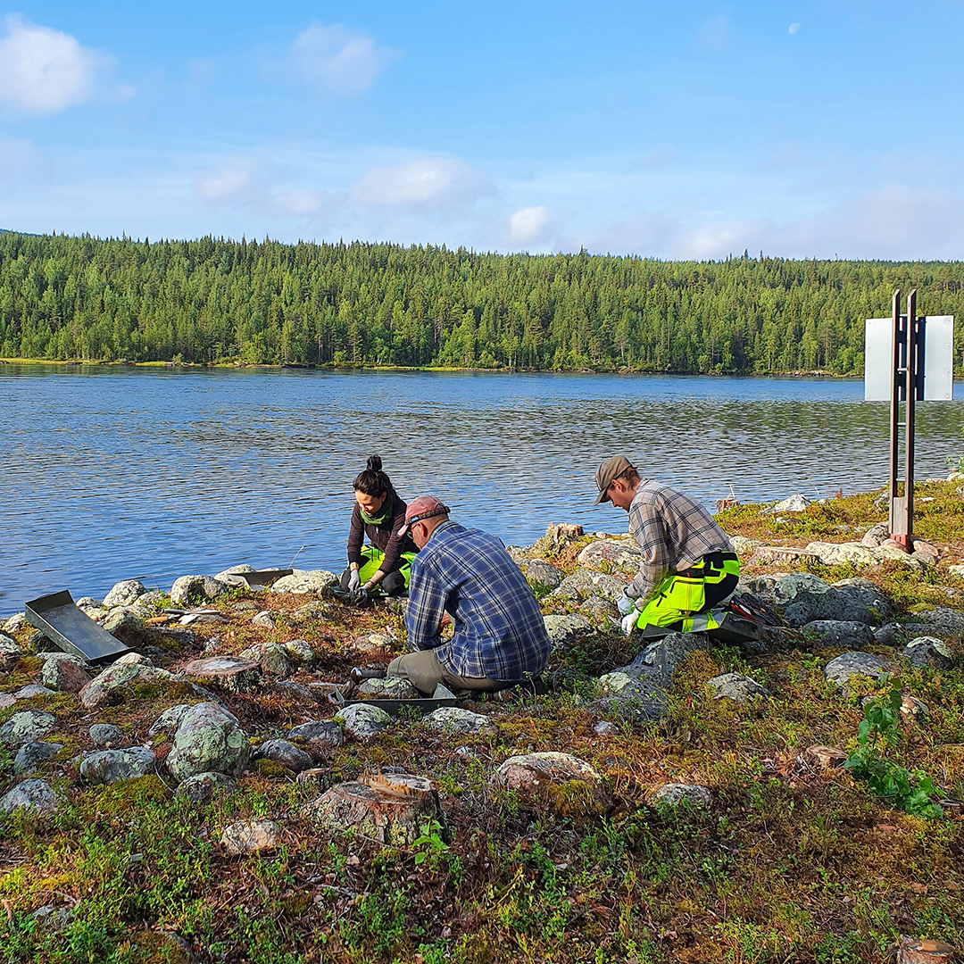 'Tre arkeologer som undersöker en stenig mark intill en älv.'